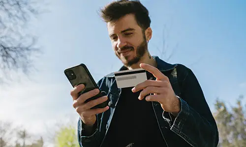 Man standing outside using cell phone in one hand and holding credit card in the other.