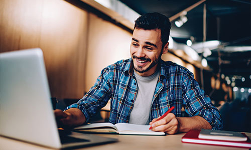 Man in library sitting at table happily looking at laptop with pen and pencil in other hand.