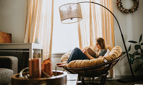 Young woman sitting in papasan chair writing in notebook with large curved lamp above her head and large window with curtains behind her.